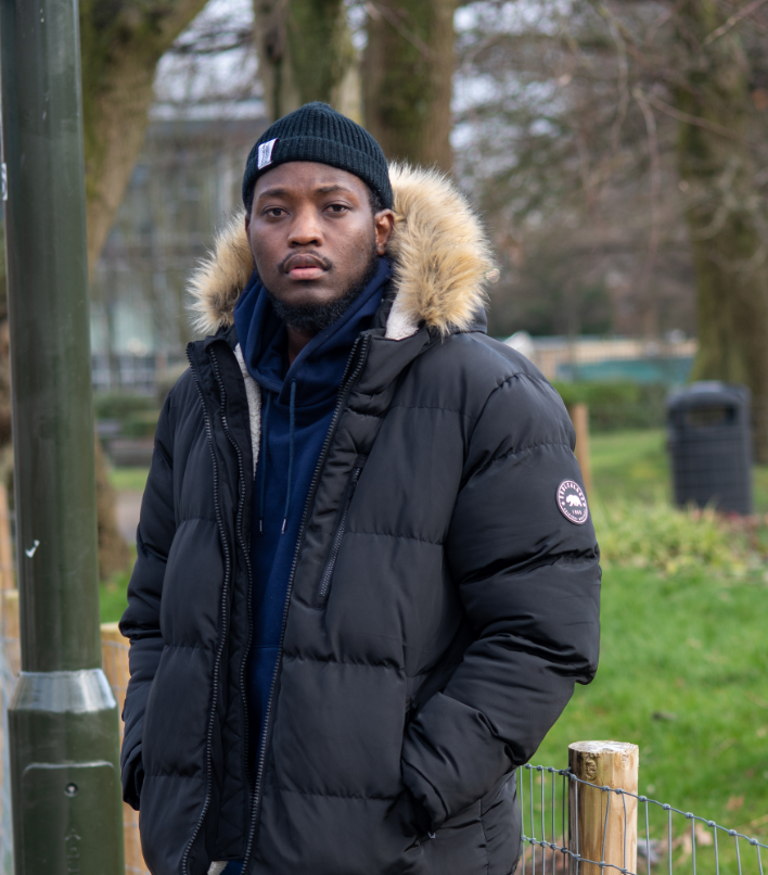 a young black man in winter clothes stands in a park, looking at the camera